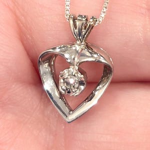 May include: A silver necklace with a heart-shaped pendant. The pendant features a small, round diamond set in the center. The chain is a delicate box chain style. The necklace is displayed against a neutral background.