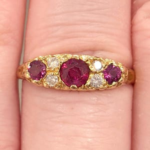 May include: A gold ring featuring a central, large, round ruby, flanked by two smaller rubies and four smaller, round, clear gemstones. The ring is set on a gold band.