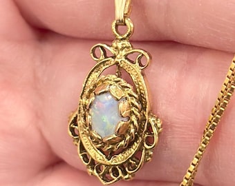 May include: A gold pendant necklace featuring an oval-shaped opal gemstone set in a decorative, filigree-style gold frame. The pendant hangs from a delicate gold box chain, showcasing a polished finish and intricate metalwork details.