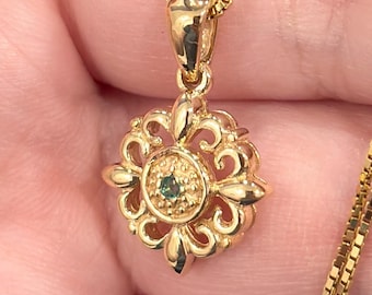 May include: A gold-toned necklace featuring a delicate box chain and a circular pendant. The pendant has an intricate openwork filigree design with a small, round, dark green gemstone set in the textured center of the piece.