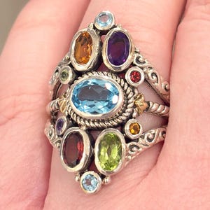 May include: A silver ring featuring an array of colorful gemstones. The ring has a large oval-shaped light blue stone at the center, surrounded by smaller stones in shades of orange, purple, green, and red. The ring's band has an ornate, textured design.