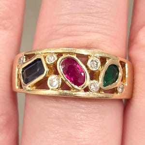 May include: A gold ring featuring a rectangular dark blue gemstone, a ruby, and an emerald, along with several small diamonds. The ring has an openwork design and is set on a finger.