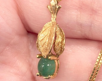 May include: A gold-colored necklace featuring a pendant with a green gemstone. The pendant has a leaf design above the gemstone. The necklace chain is a thin, gold-colored box chain. The gemstone is a round, polished, green stone.