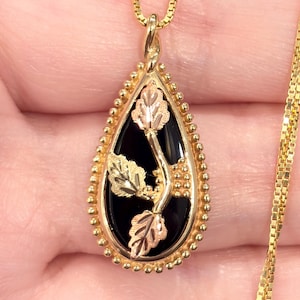 May include: A gold-toned necklace with a teardrop pendant. The pendant features a black center with gold and rose-gold leaf and vine details. The pendant is framed with a beaded border. The chain is a delicate box chain.