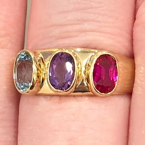 May include: A gold ring featuring three oval gemstones: a light blue aquamarine, a purple amethyst, and a vibrant red ruby. The ring's band is a polished gold, showcasing the colorful gemstones.