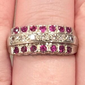 May include: A silver-toned ring featuring three horizontal rows of stones. The top and bottom rows consist of small, round, vibrant pink rubies, while the center row contains a series of clear, sparkling white diamonds set in a metallic band.