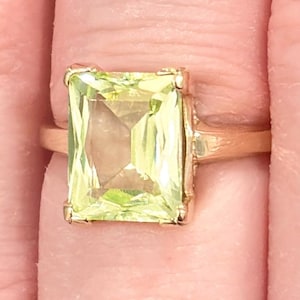 May include: A gold ring featuring a large, rectangular, light green gemstone. The gemstone is set in a gold band, and the ring is shown on a finger. The ring's design is simple, with a focus on the gemstone's size and color.