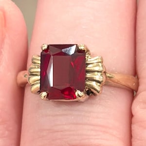 Peut inclure: Une bague dorée ornée d'une grande pierre précieuse rouge foncé de forme rectangulaire. La pierre est sertie dans une monture décorative avec un motif nervuré de chaque côté. L'anneau est en or poli.