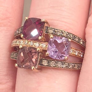 May include: Three gold rings, each with a large, square-cut gemstone in shades of purple, pink, and brown. The bands are decorated with smaller stones. The rings are stacked on a finger, highlighting their design and the contrast of colours.