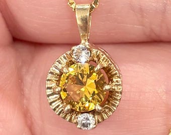 May include: A gold pendant necklace featuring a round, yellow gemstone surrounded by a halo of smaller white gemstones. The pendant hangs from a delicate gold chain. The necklace is displayed against a skin-toned background.