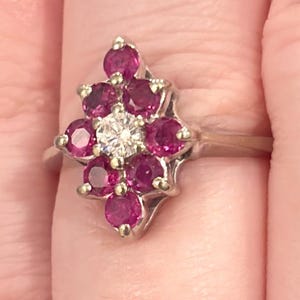 May include: A silver-toned ring featuring a cluster of eight round, vibrant pink stones arranged in a marquise-shaped pattern, with a single clear, round-cut stone centered in the middle of the cluster, set on a plain, polished metal band.
