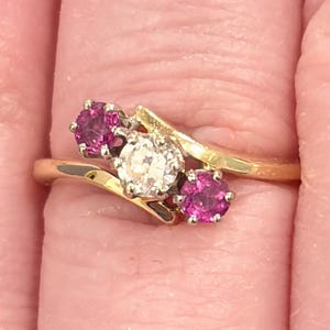 May include: A gold ring featuring a central, round, clear gemstone flanked by two smaller, round, pink gemstones. The ring's band curves gracefully, creating a stylish and elegant design. The gemstones are set in a gold band.