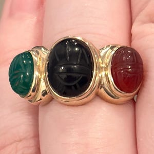 May include: A gold ring featuring three oval-shaped scarab-style carved gemstones. The center stone is black, flanked by a green stone on the left and a red stone on the right. Each gemstone is set in a polished gold band with a smooth, rounded finish.