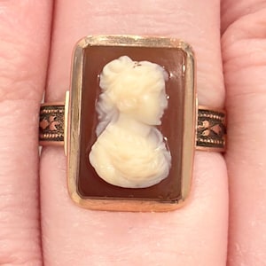 May include: A gold-toned ring featuring a rectangular cameo with a profile of a woman. The cameo is cream-colored against a brown background. The ring band has a decorative pattern.
