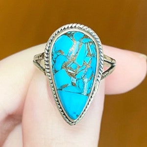 May include: A silver ring featuring a teardrop-shaped turquoise stone with a copper matrix. The stone is set in a silver bezel with a twisted rope design. The ring has a delicate band with decorative cutouts.