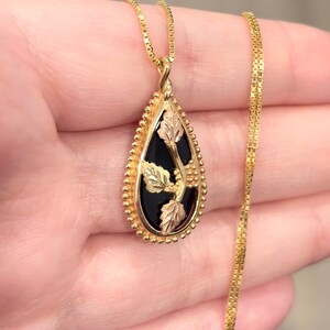May include: Gold-tone pendant necklace with a teardrop-shaped black onyx stone. The pendant features gold and rose-gold leaf accents. The necklace chain is a delicate gold color.
