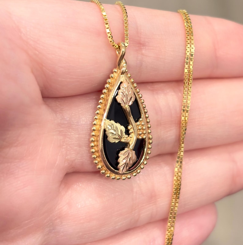 May include: Gold-tone pendant necklace with a teardrop-shaped black onyx stone. The pendant features gold and rose-gold leaf accents. The necklace chain is a delicate gold color.