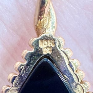 May include: Close-up of a gold-colored pendant with a triangular black stone. The pendant has a decorative, textured border and a loop at the top. The inscription "10K" is visible on the pendant.