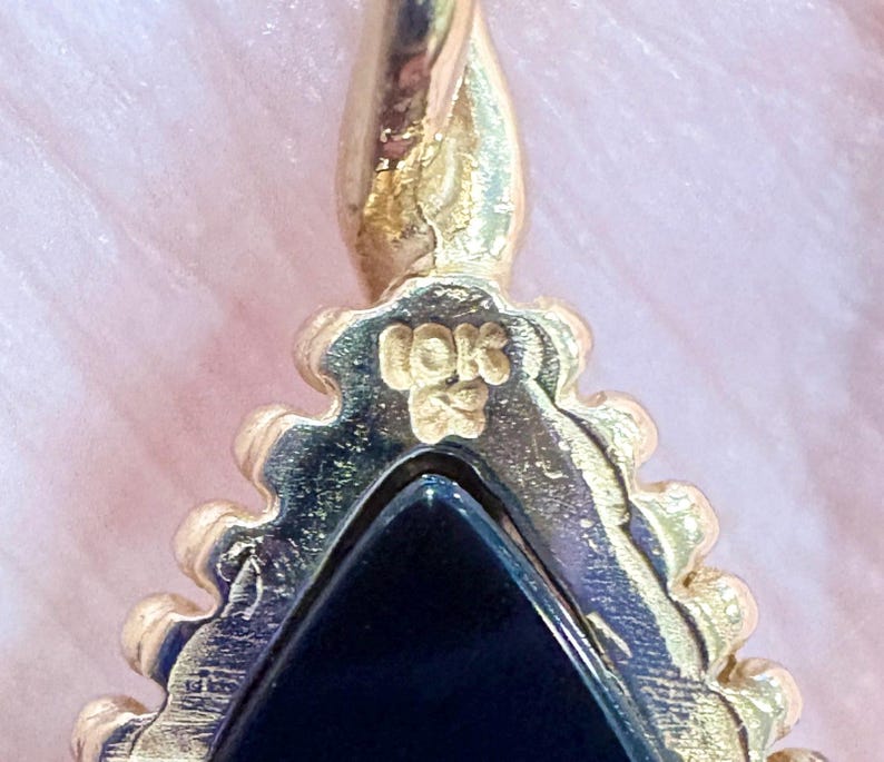 May include: Close-up of a gold-colored pendant with a triangular black stone. The pendant has a decorative, textured border and a loop at the top. The inscription "10K" is visible on the pendant.