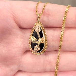 May include: Gold-tone necklace with a teardrop pendant featuring a black onyx center and gold leaf accents. The pendant is framed with a beaded border. The chain is a delicate box chain.