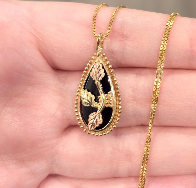 May include: Gold-tone necklace with a teardrop pendant featuring a black onyx center and gold leaf accents. The pendant is framed with a beaded border. The chain is a delicate box chain.