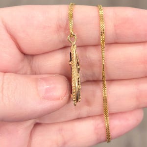 May include: A gold-colored necklace with a pendant. The pendant has an ornate design with a central shape and decorative elements. The chain is a fine, box-style chain. The necklace is held in a hand.