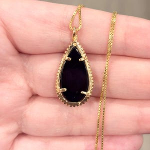 May include: A gold-colored necklace featuring a teardrop-shaped pendant with a black stone center. The pendant is framed by a detailed gold border and hangs from a delicate gold chain. The necklace is displayed against a neutral background.