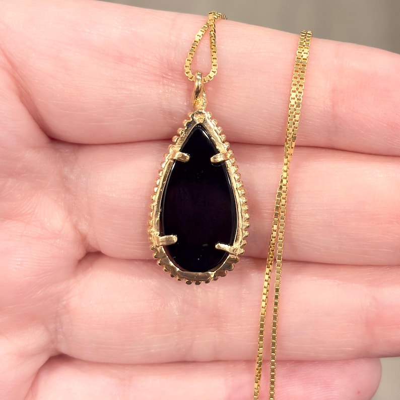 May include: A gold-colored necklace featuring a teardrop-shaped pendant with a black stone center. The pendant is framed by a detailed gold border and hangs from a delicate gold chain. The necklace is displayed against a neutral background.