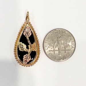 May include: Gold-tone pendant with a black teardrop-shaped center, featuring a floral design with gold, rose gold, and green accents. The pendant is next to a U.S. dime for size comparison. The pendant has a beaded border.