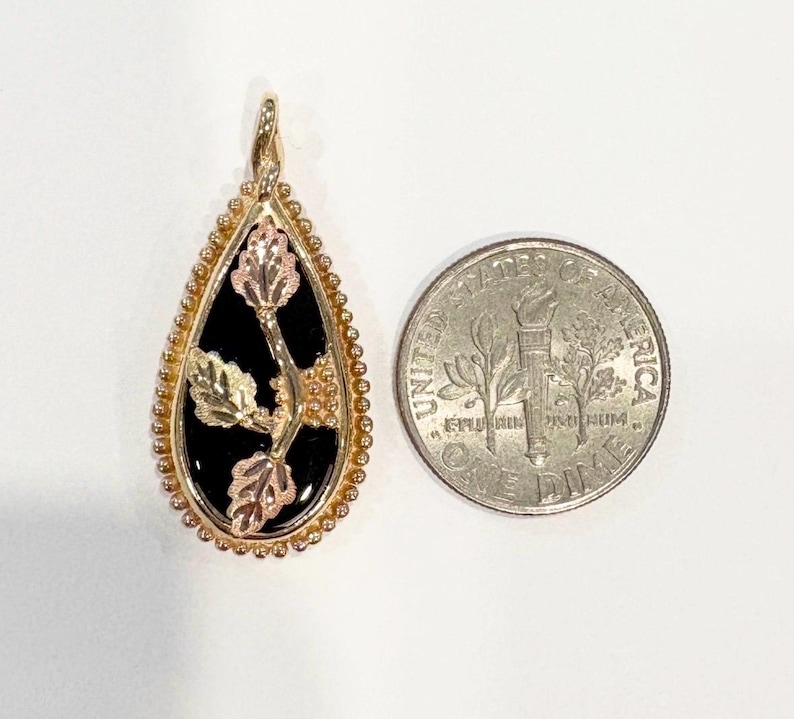 May include: Gold-tone pendant with a black teardrop-shaped center, featuring a floral design with gold, rose gold, and green accents. The pendant is next to a U.S. dime for size comparison. The pendant has a beaded border.