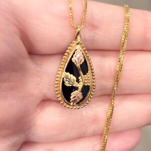 May include: A gold-toned pendant necklace with a black teardrop-shaped center. The pendant features gold and rose-gold leaf accents. The necklace chain is a delicate gold box chain.