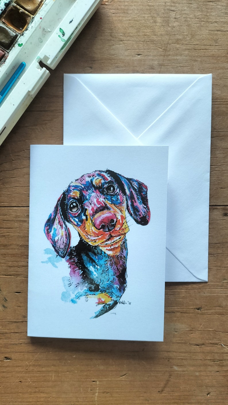 Op de afbeelding: Een kleurrijk aquarel schilderij van een teckel met grote ogen en een speelse uitdrukking. De hond is geschilderd in tinten blauw, roze, geel en zwart.