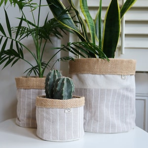 May include: Three fabric plant pots with a burlap trim. The pots are white with grey stripes and the text "hihi" is printed on the front of each pot. The pots are filled with plants, including a cactus and a snake plant.