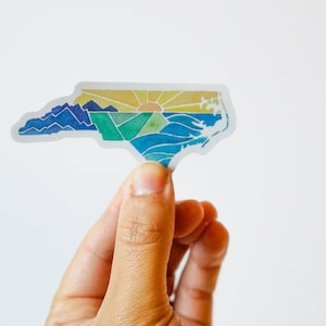 May include: A sticker of the state of North Carolina with a watercolor design of mountains, a sunset, and the ocean.