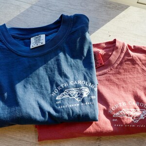 K&ouml;nnte beinhalten: Zwei T-Shirts, eines in Marineblau und eines in Rotbraun, werden gezeigt. Jedes Shirt hat eine wei&szlig;e Grafik der Umrisse von North Carolina, den Text "NORTH CAROLINA", das Jahr "1789" und den Satz "esse quam videri".