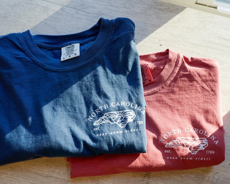 K&ouml;nnte beinhalten: Zwei T-Shirts, eines in Marineblau und eines in Rotbraun, werden gezeigt. Jedes Shirt hat eine wei&szlig;e Grafik der Umrisse von North Carolina, den Text "NORTH CAROLINA", das Jahr "1789" und den Satz "esse quam videri".