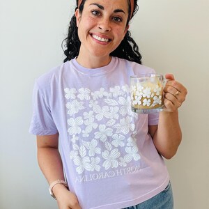 Op de afbeelding: Een vrouw draagt een lavendelkleurig T-shirt met een wit bloemenontwerp en de tekst "NORTH CAROLINA" gedrukt ondersteboven. Ze houdt een witte mok vast met een bloemenontwerp en een bruine drank erin.