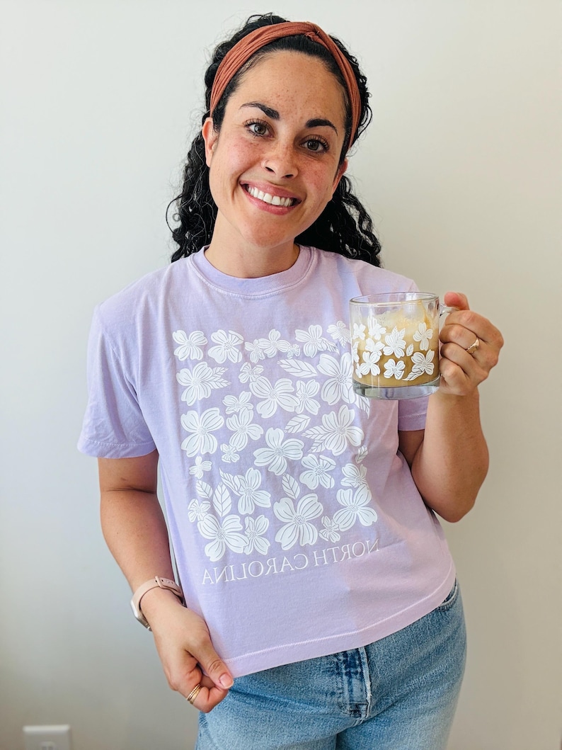 Op de afbeelding: Een vrouw draagt een lavendelkleurig T-shirt met een wit bloemenontwerp en de tekst "NORTH CAROLINA" gedrukt ondersteboven. Ze houdt een witte mok vast met een bloemenontwerp en een bruine drank erin.