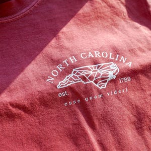 K&ouml;nnte beinhalten: Ein weinrotes T-Shirt mit einer wei&szlig;en Grafik der Umrisse des Bundesstaates North Carolina, den Worten "NORTH CAROLINA" dar&uuml;ber und "1789" darunter. Der Satz "esse quam videri" ist darunter gedruckt.