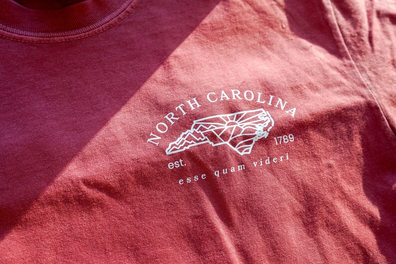 K&ouml;nnte beinhalten: Ein weinrotes T-Shirt mit einer wei&szlig;en Grafik der Umrisse des Bundesstaates North Carolina, den Worten "NORTH CAROLINA" dar&uuml;ber und "1789" darunter. Der Satz "esse quam videri" ist darunter gedruckt.