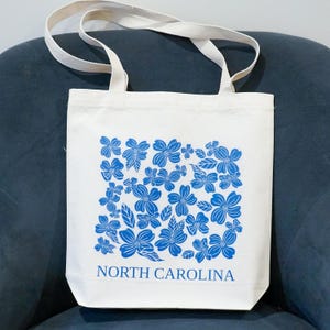 May include: A white canvas tote bag with blue floral print and the text "NORTH CAROLINA".
