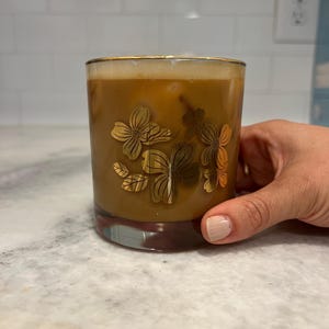 May include: A glass of iced coffee in a clear glass with a gold floral design. The glass is being held by a hand.