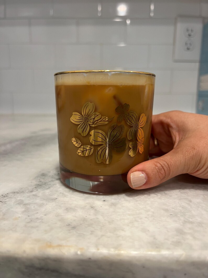 May include: A glass of iced coffee in a clear glass with a gold floral design. The glass is being held by a hand.