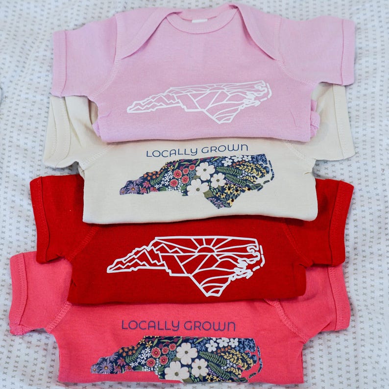 May include: Four baby onesies stacked on top of each other. The onesies are pink, beige, red, and pink. Each onesie features a white outline of the state of North Carolina with a different design inside. The top onesie has a geometric design, the second onesie has a floral design, the third onesie has a mountain and sun design, and the bottom onesie has a floral design. Each onesie also has the text "Locally Grown" printed on it.