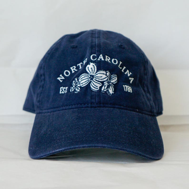 May include: A navy blue baseball cap with white embroidery. The embroidery reads "North Carolina Est 1789" and features a design of three white dogwood flowers.