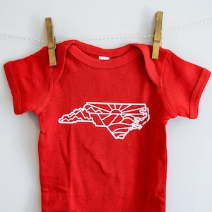 May include: Red baby onesie with a white outline of the state of North Carolina, featuring mountains and a sunrise.