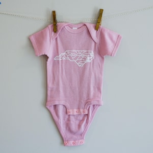 May include: A pink baby onesie with a white outline of the state of North Carolina, featuring mountains and a sun graphic.