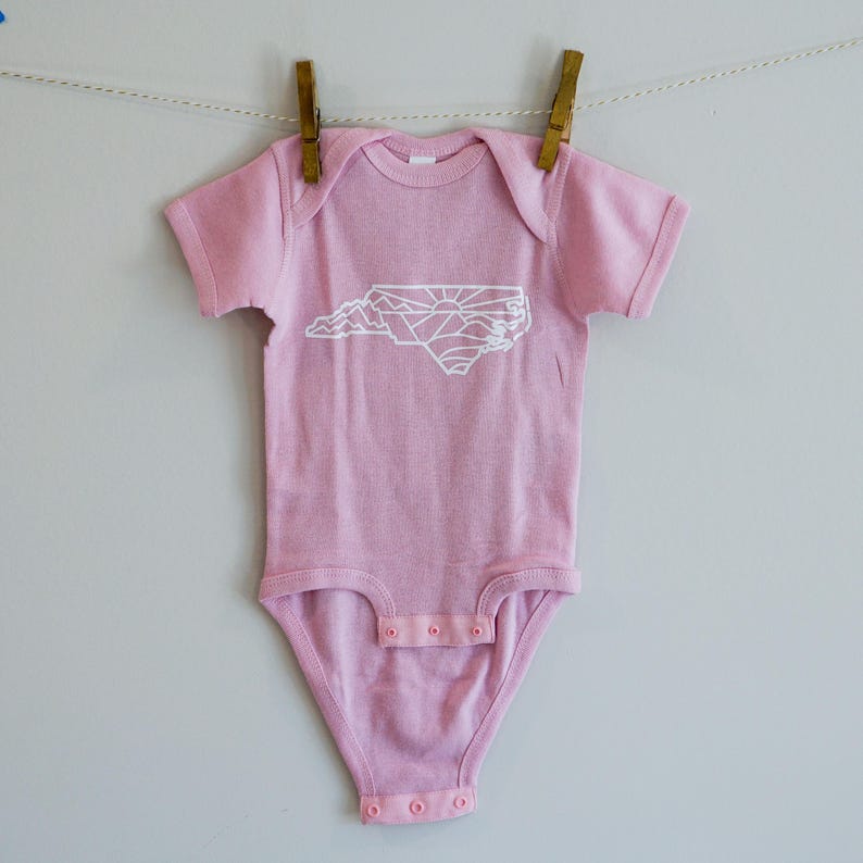 May include: A pink baby onesie with a white outline of the state of North Carolina, featuring mountains and a sun graphic.