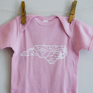 May include: A pink baby onesie with a white outline of the state of North Carolina, featuring a sunrise and mountains inside the state shape.