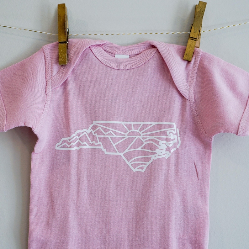 May include: A pink baby onesie with a white outline of the state of North Carolina, featuring a sunrise and mountains inside the state shape.